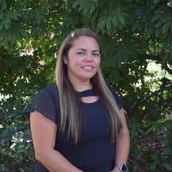 Dominica Albitre; A woman with long hair, wearing a black blouse, stands in front of lush green foliage, smiling softly.