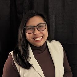 Hapy Ting; A smiling woman with long black hair wearing glasses, a brown turtleneck, and a white vest, against a black background.