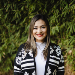 Monica Cardenas, wearing a black and white patterned cardigan with a white long sleeve stands outdoors in front of bamboo leaves