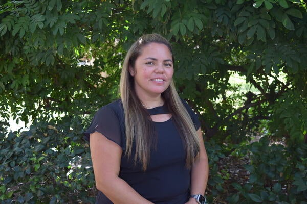 Dominica Albitre; A woman with long hair, wearing a black blouse, stands in front of lush green foliage, smiling softly.
