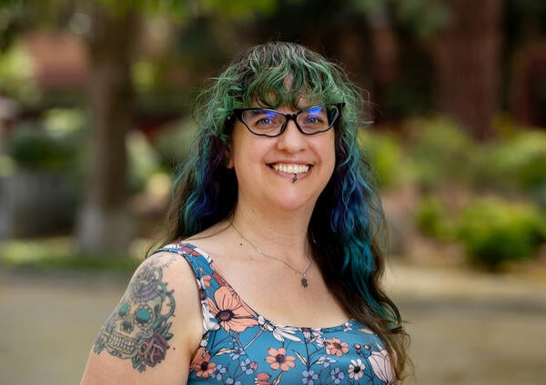 Cierra Covarrubias; Smiling woman with long, wavy green and blue hair, wearing glasses and a floral top. Her tattooed arm is visible. Background is a blurred garden.