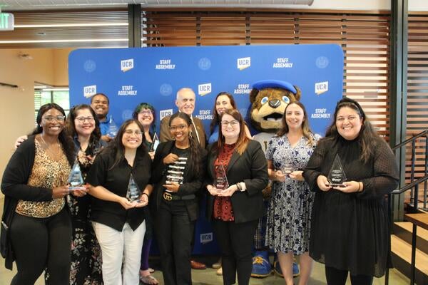 2024-2025 Staff Assembly Board, a group of 12 holding awards in front of a Staff Assembly branded backdrop