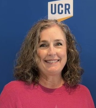 Kelly Jones; Woman with curly hair smiles, standing in front of a blue background with the UCR logo. She wears a pink shirt, conveying a friendly and professional tone.