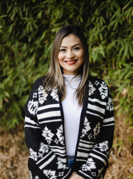 Monica Cardenas, wearing a black and white patterned cardigan with a white long sleeve stands outdoors in front of bamboo leaves