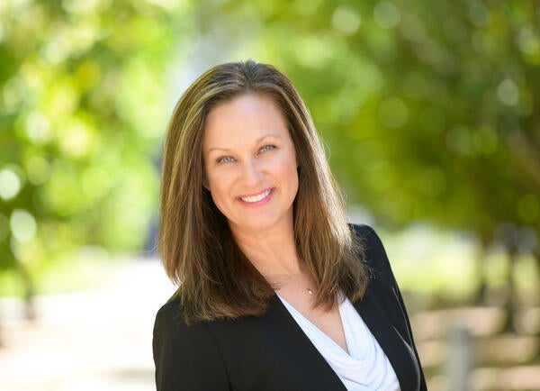 headshot of a JoAnna Van Brocklin  with light brown highlighted hair that comes to her shoulders. She is smiling.he wears a white blouse with a cowl neckline and a small silver necklace. She has on a black blazer. Her arms are down and not visible in the frame, but she seems to be standing in front of greenery that is in a park, blurred, due to a shallow depth of field.